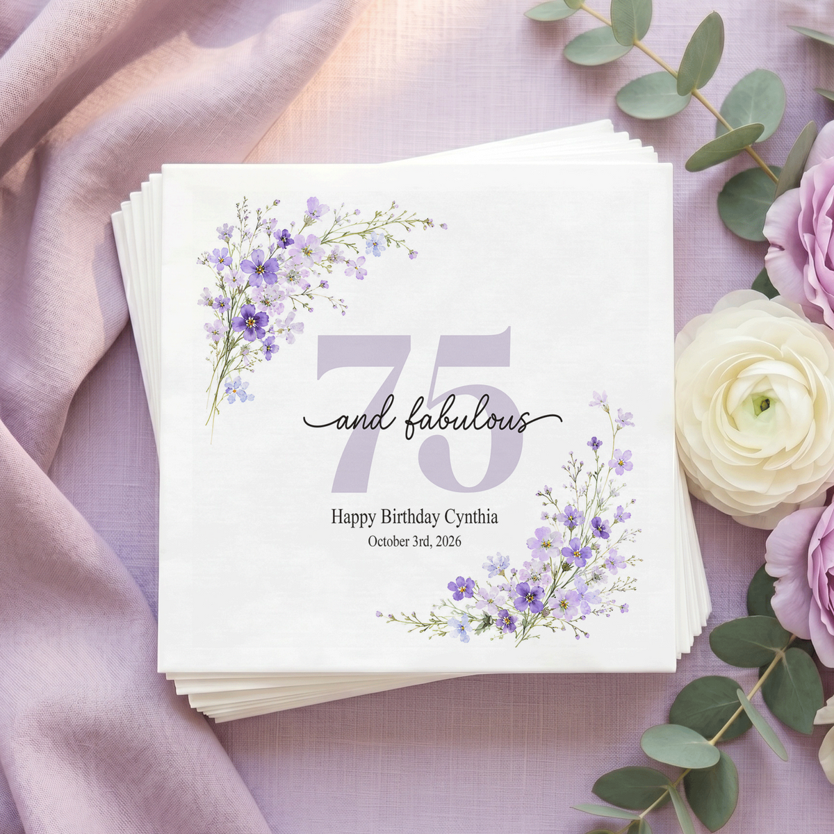 White napkin with a pale lilac 75, black “and fabulous” script, purple wildflowers in opposite corners, and a personalized message and date.