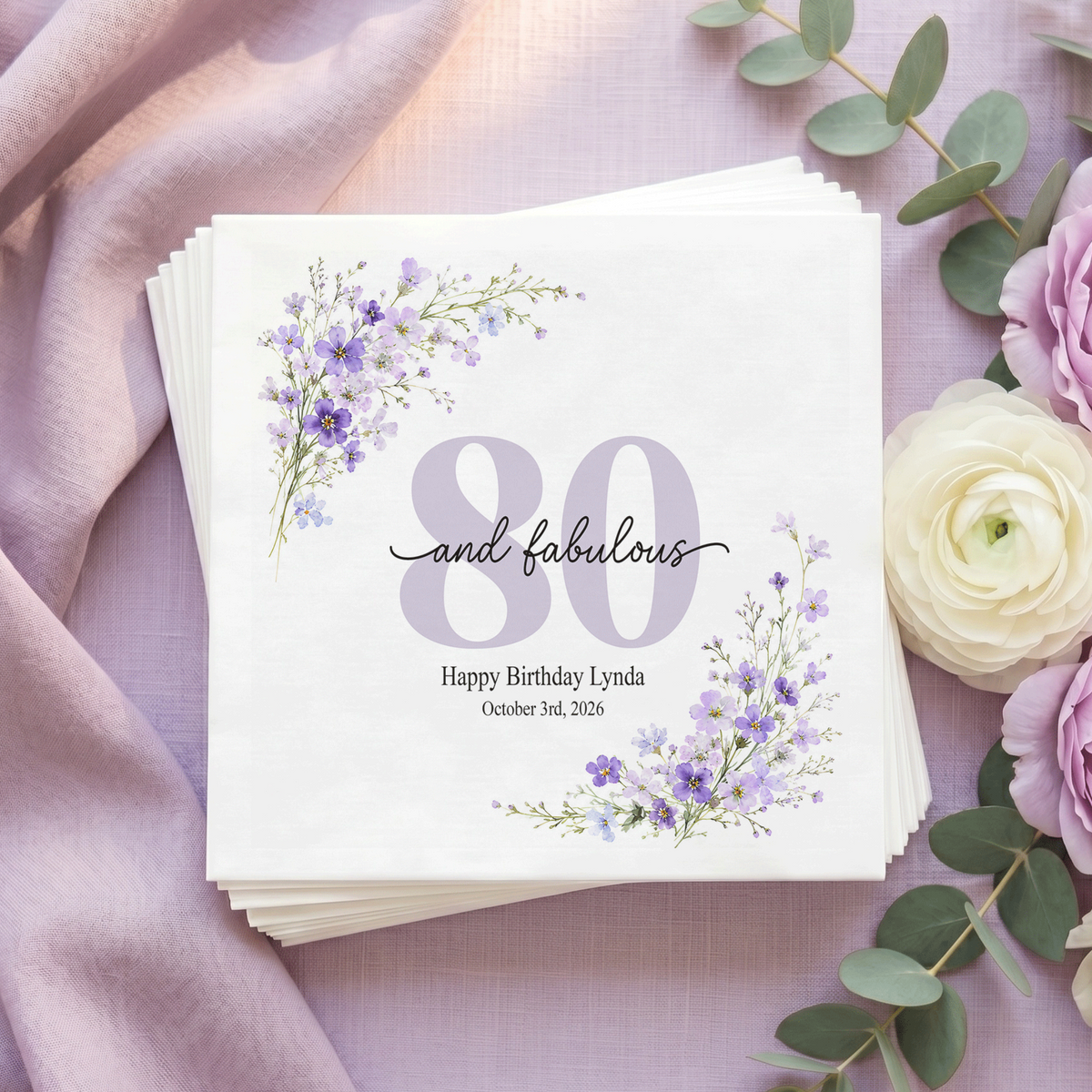 White napkin with a pale lilac 80, black “and fabulous” script, purple wildflowers in opposite corners, and a personalized message and date.

