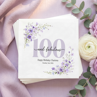 White napkin with a pale lilac 100, black “and fabulous” script, purple wildflowers in opposite corners, and a personalized message and date.