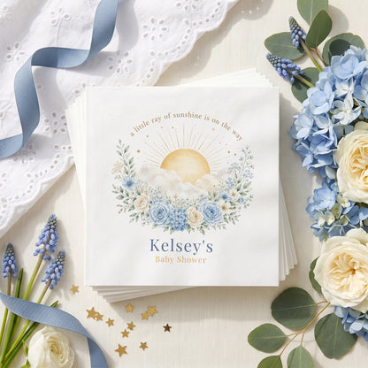 Dusty blue baby shower napkin with a gold rayed sun, white clouds, blue roses and a personalized name in deep blue